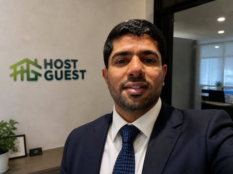 Man in dark suit and tie smiling at camera in office with Host Guest logo on wall behind him