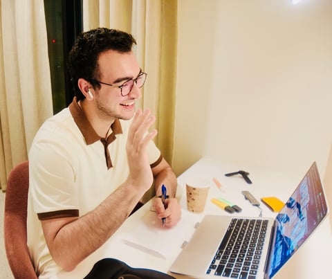 Man wearing glasses and beige shirt sitting at desk with laptop, smiling while working on a drawing or design project
