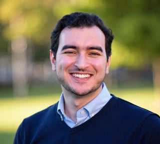 Smiling man with dark hair wearing a navy sweater over a light blue shirt, outdoors with green background