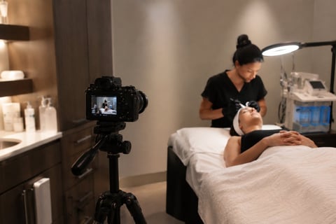 Esthetician performing facial treatment on client while being filmed by camera on tripod in spa setting