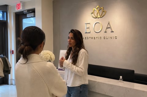 Two women at reception desk of EOA Aesthetic Clinic, one facing away and one behind counter wearing white shirt