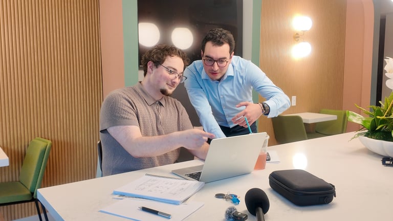Two men working together at a desk, one in gray shirt pointing at laptop screen while the other in blue shirt leans in to look, with office supplies and plants nearby
