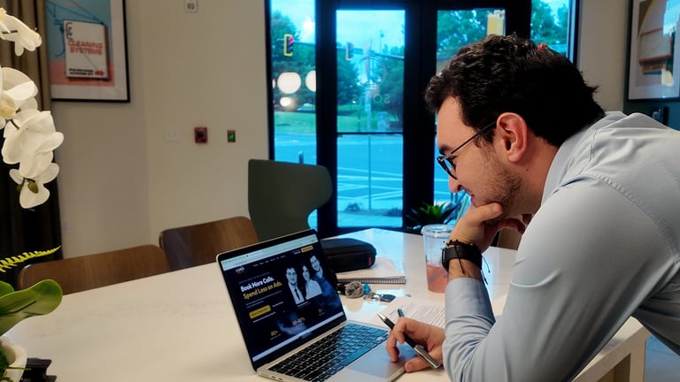 Man wearing glasses leaning over a laptop at a desk, working with a document and pen in a modern office setting with blue-tinted windows